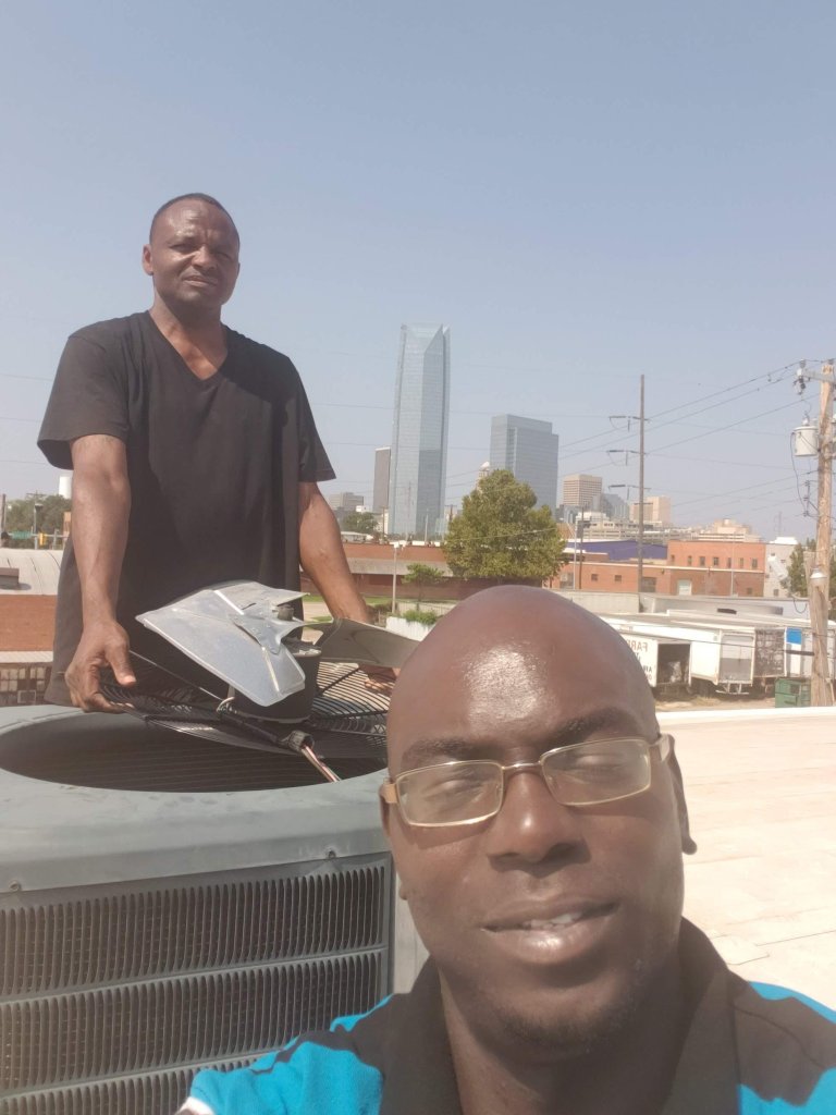 Two technicians on a flat roof with a rooftop AC unit and its fan assembly, Oklahoma City skyline behind