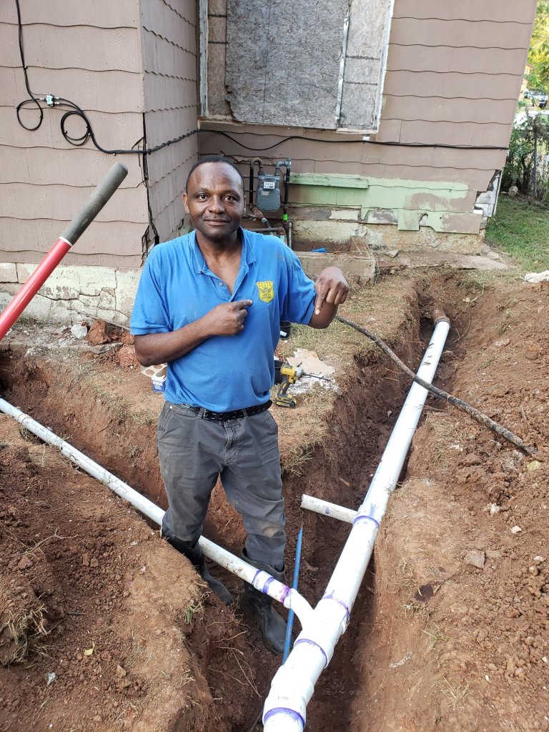 Team member in a branded blue polo standing in a trench beside new white PVC drain pipe