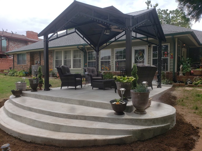 Curved three-step concrete patio with a black metal pergola and patio furniture behind a house